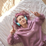Happy funny teen girl with pink hair wear headphones lying in comfortable bed listening new pop music enjoying singing song with eyes closed relaxing in cozy bedroom at home. Top view from above.