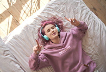 Happy funny teen girl with pink hair wear headphones lying in comfortable bed listening new pop music enjoying singing song with eyes closed relaxing in cozy bedroom at home. Top view from above.