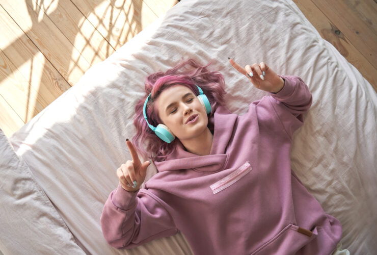 Happy funny teen girl with pink hair wear headphones lying in comfortable bed listening new pop music enjoying singing song with eyes closed relaxing in cozy bedroom at home. Top view from above.