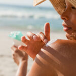 Young woman applying sunscreen on her shoulder while sitting on the beach