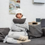 freezin young woman in warm clothes sitting on couch and hugging cushion at home. Learn about cold flashes and menopause.