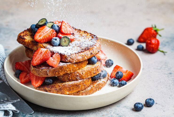 pile of french toast on a white plate with strawberries, blueberries, and powdered sugar