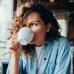 a woman with curly brown hair drinks a beverage out of a coffee cup