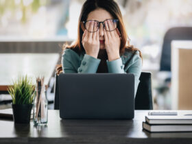 Stressed out woman with brown hair and teal sweater rubbing her eyes while sitting at a laptop computer.