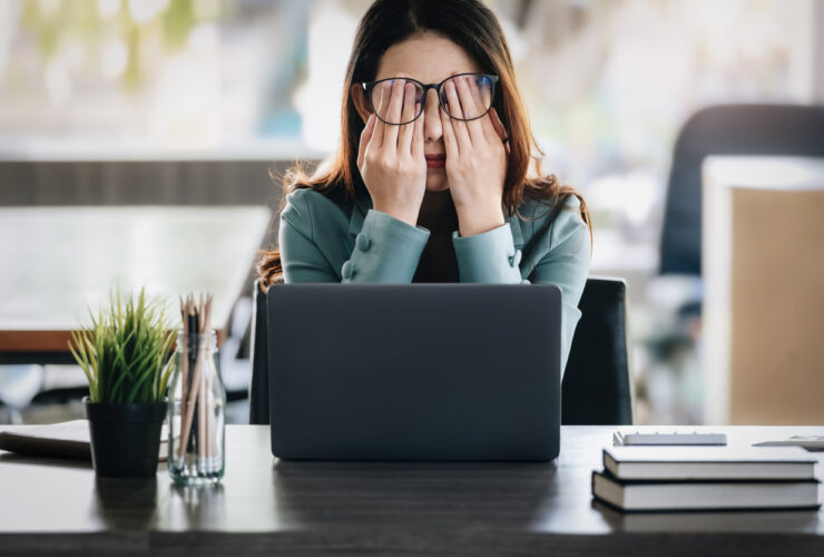 Stressed out woman with brown hair and teal sweater rubbing her eyes while sitting at a laptop computer.