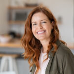 Beautiful woman in her 40s with red hair and a green jacket smiling. Learn when to take progesterone for perimenopause at GloWellmag.com